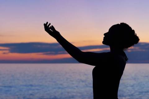A photo of a woman in silhouette with her hands lifted against the background of the ocean at sunset